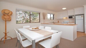 A modern kitchen with white cabinetry, a central island, and a dining area with a white table and chairs. - Promenade 11, 8 Intrepid Close - air conditioned unit with beautiful water views (Nelson Bay)