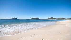 A beach with clear blue water, sandy shore, and distant hills. - Promenade 11, 8 Intrepid Close - air conditioned unit with beautiful water views (Nelson Bay)