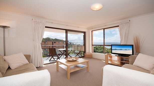 A modern living room with a large TV, comfortable seating, a balcony with a view, and a large window. - Promenade 11, 8 Intrepid Close - air conditioned unit with beautiful water views (Nelson Bay)