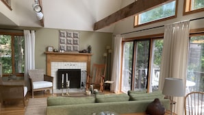 Una sala de estar acogedora con chimenea, sofá verde, muebles de madera y ventanas amplias que ofrecen vistas al exterior.