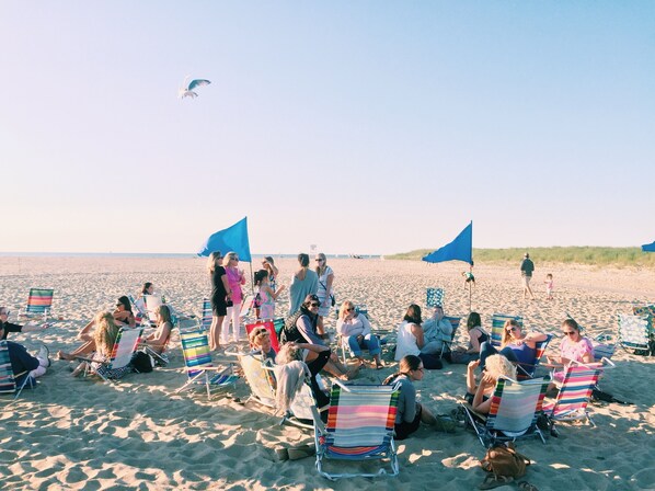 Une scène de plage où l’on voit des personnes assises sur des chaises de plage, à l’ombre de parasols bleus, d’autres debout, et un goéland qui vole dans le ciel.