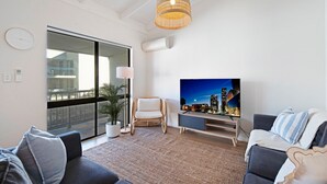 A modern living room with a large TV, wicker chair, and a view of the city at night through a sliding door.