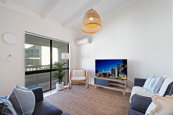 A modern living room with a large TV, wicker chair, and a view of the city at night through a sliding door.