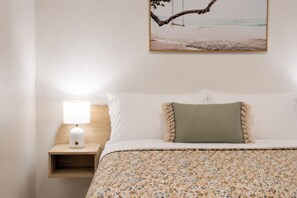 Un letto rifatto con cura, con una coperta a motivi floreali, un cuscino verde e un cuscino bianco. Un comodino in legno con una lampada bianca si trova alla sinistra.