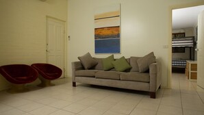 Uma sala de estar com sofá, duas cadeiras vermelhas, um quadro na parede e uma porta que dá para outro cômodo.