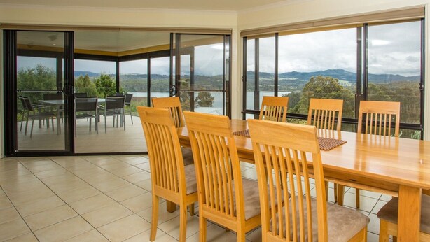 Uma área de jantar com uma mesa grande e cadeiras, grandes janelas com vista para a paisagem e uma porta de vidro deslizante que dá para um pátio externo.