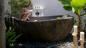 Ein Wellnessbereich mit einer großen Steinbadewanne, einem kleinen weißen Schrank mit Flaschen und einer großen Grünpflanze.