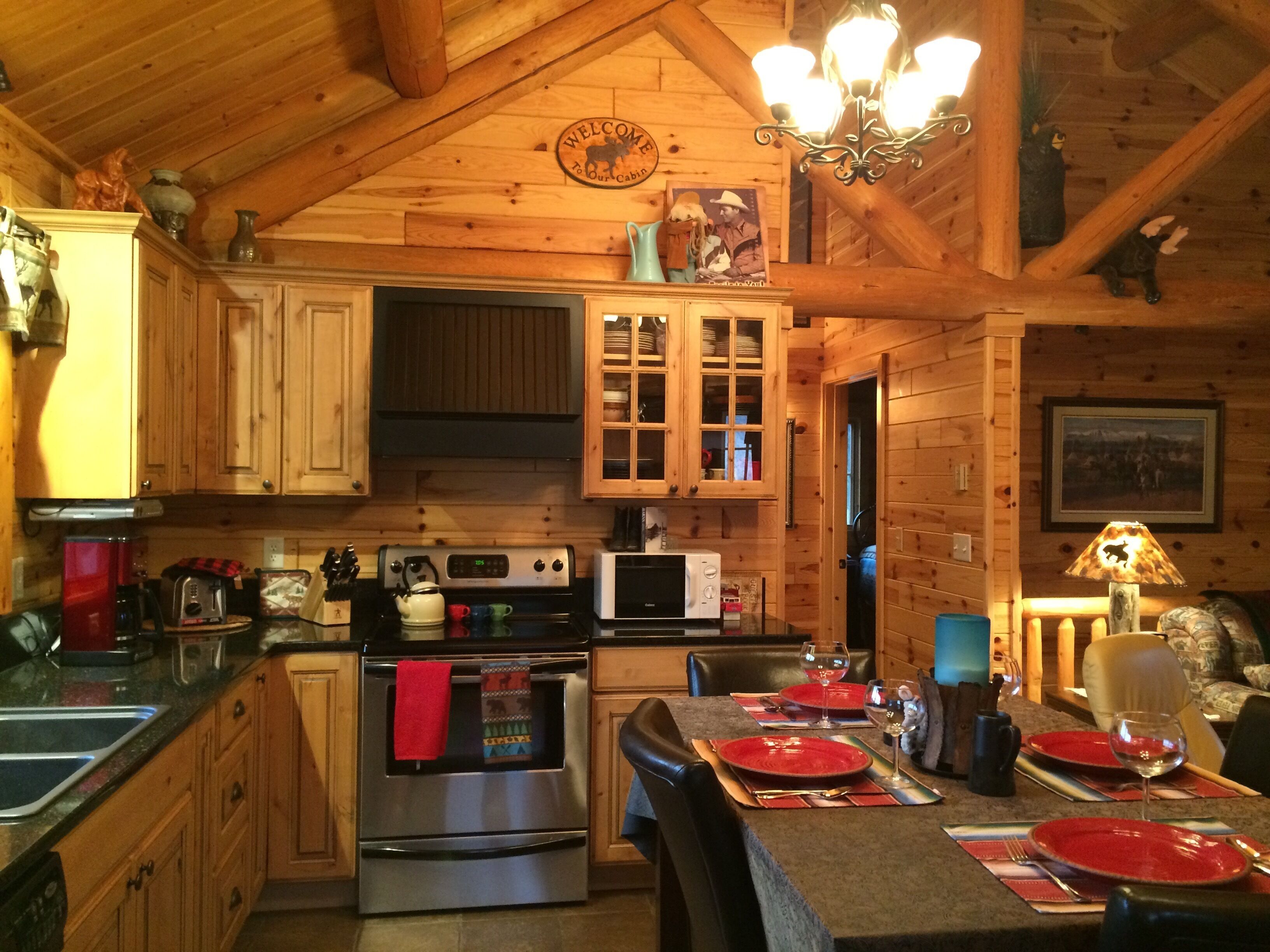 Une cuisine de style rustique, aménagée comme un chalet, avec des meubles en bois, une cuisinière, un four à micro-ondes et une table à manger pour deux personnes, agrémentée de sets de table rouges et de verres à vin.