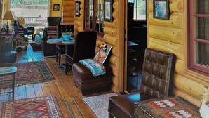 Interior de una cabaña de troncos con techo alto, paredes de madera y un ventanal grande. Hay varias zonas de estar con sillones de cuero y un sofá.