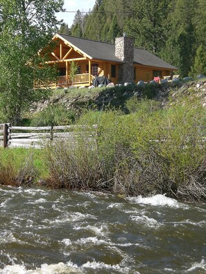 Una baita di legno con camino in pietra, circondata da fitta vegetazione e un fiume.