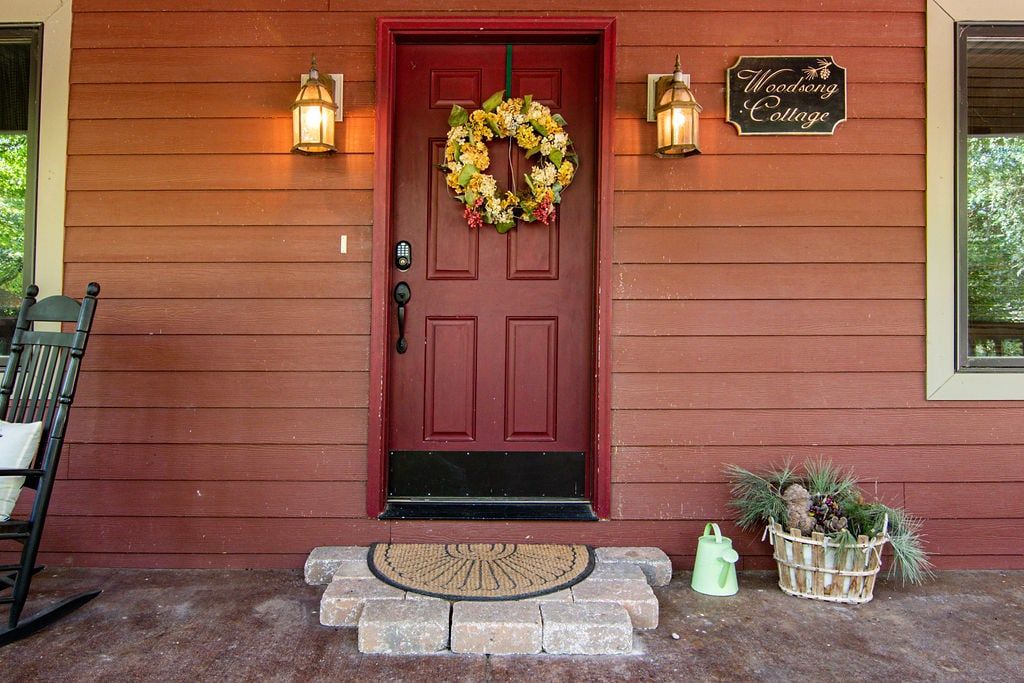 A red door with a wreath, two wall-mounted lights, a sign reading 'Woodsong Cottage,' a bench, and a welcome mat.