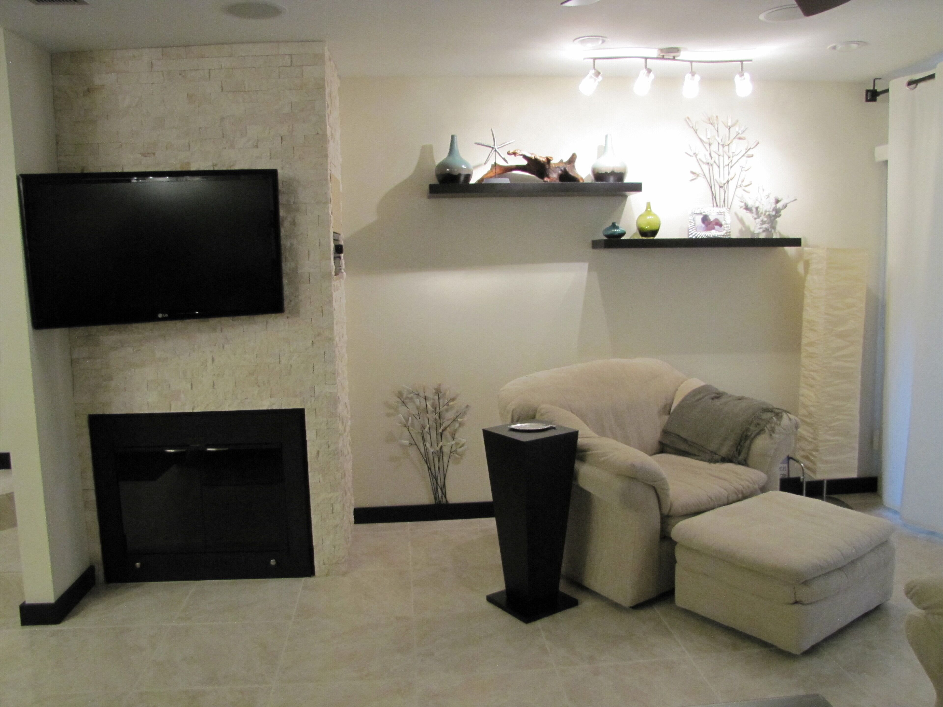 A modern living room with a flat-screen TV mounted on a brick wall, a fireplace, a beige sofa, and decorative shelves with vases.