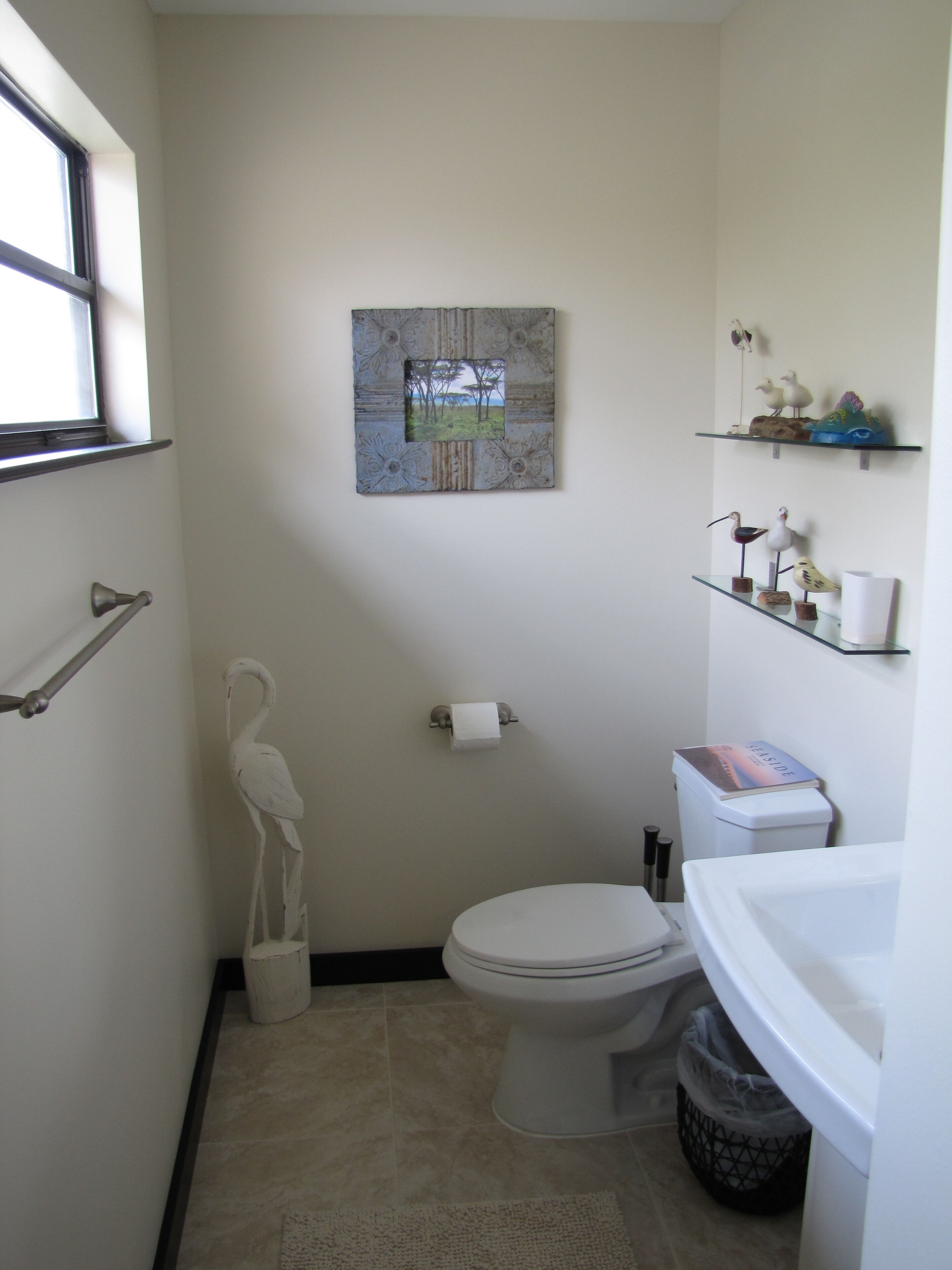 A small bathroom with a toilet, a sink, a mirror, a towel rack, a trash bin, and a framed picture on the wall.