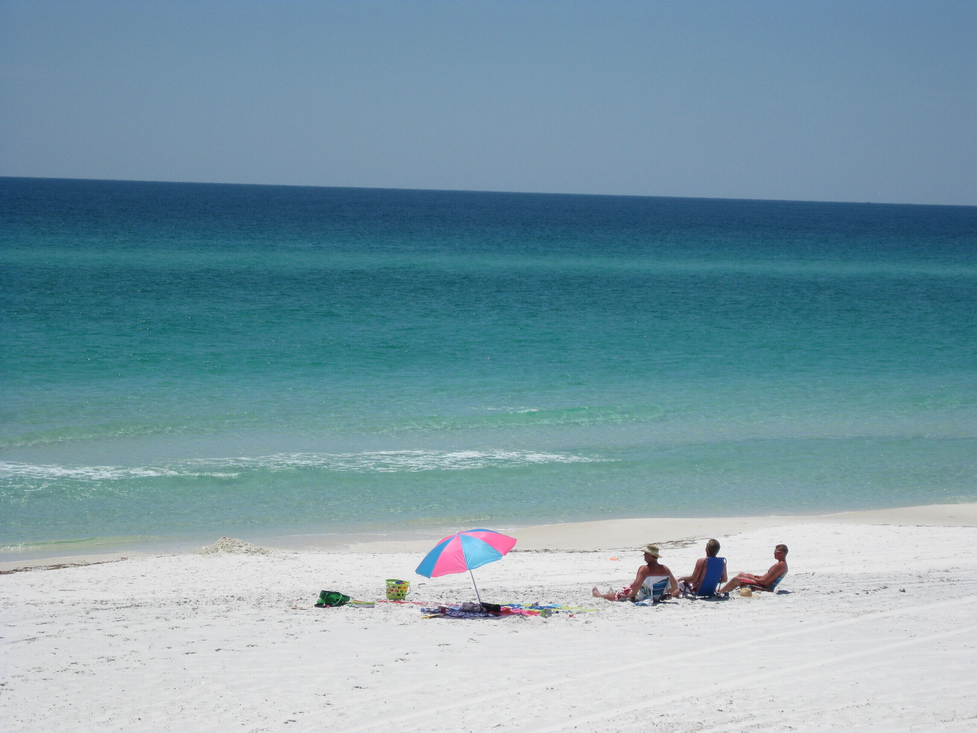 A beach scene with a clear sky, calm sea, and three people under a colorful umbrella.