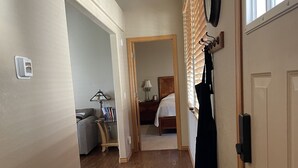 A hallway with wooden flooring, a coat rack, and a door leading to a bedroom with a bed and a nightstand. - BEAUTIFUL SPACIOUS APARTMENT- 1 Bedroom with Living Room & Kitchen. Sleeps 4. (Monument)