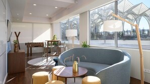 Una sala moderna con un sofá curvo, una mesa de centro redonda y dos sillones. Hay una zona de comedor con una mesa y sillas, y un ventanal con vistas a un edificio de estructura de techo singular.