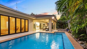Une piscine avec un patio couvert, entourée d’une végétation luxuriante, et une maison dotée de grandes fenêtres et d’un toit en tuiles.