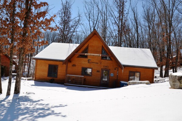 A wooden cabin with a snow-covered roof, situated in a forested area.