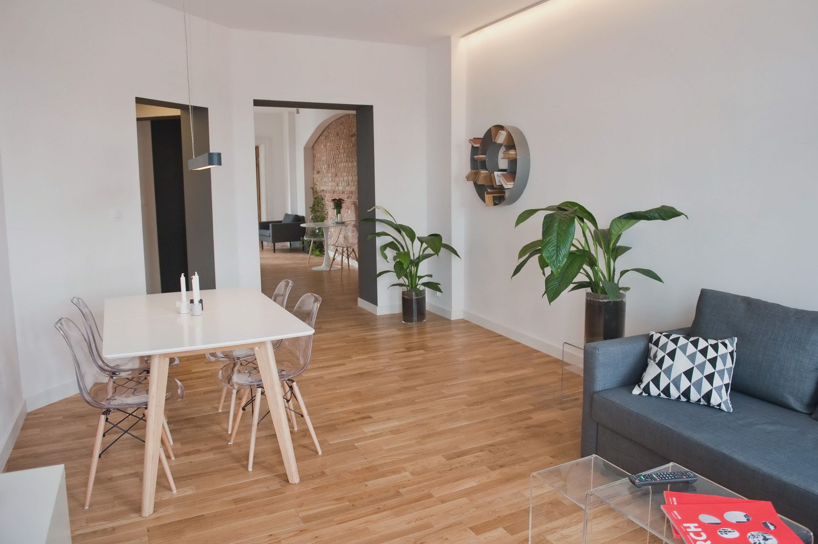 A modern living room with a dining area, featuring a sofa, a table, and chairs. There are potted plants and a wall-mounted shelf.