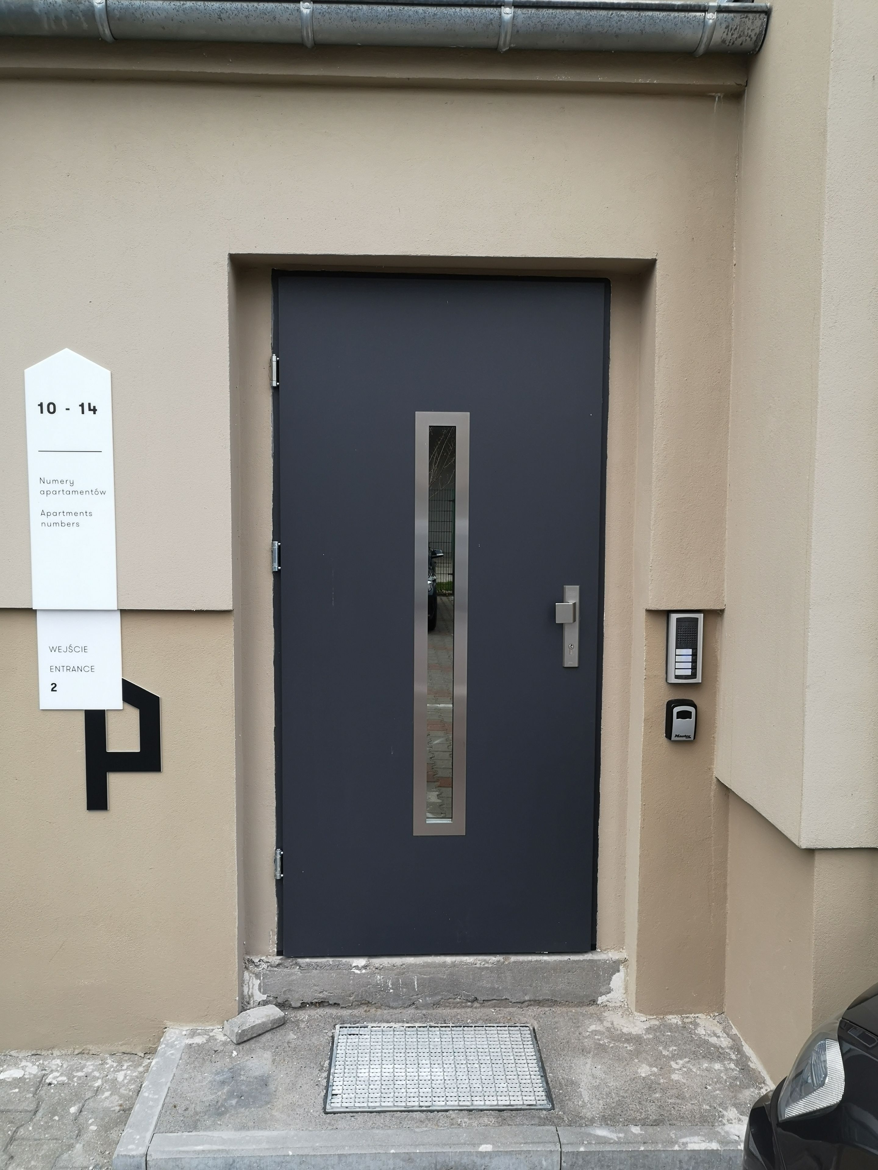 Une entrée d’immeuble moderne avec une porte sombre, une boîte aux lettres et une plaque indiquant les numéros d’appartement 10 à 14.
