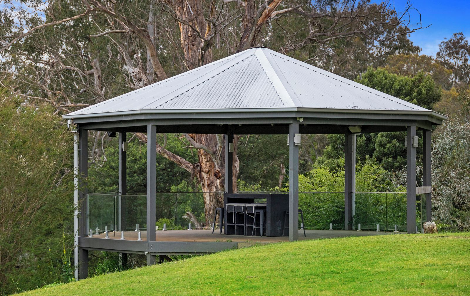 Un pavillon doté d’un toit en métal et de rampes en verre, entouré d’arbres et d’herbe.