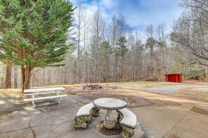 A picnic table with a stone pedestal base, surrounded by a forested area with a bench and a red structure in the background. - Spacious Group Retreat w/ Game Room in Marshall! (Marshall)