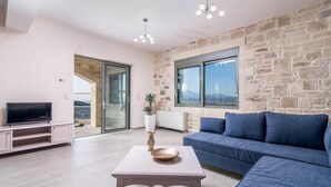 Un salon moderne avec un mur en pierre, un canapé bleu, un téléviseur posé sur un meuble et une grande fenêtre donnant sur un paysage.