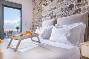 A bedroom with a stone wall, a bed with white linens, a bedside table, and a window with a view. - Villa Ovgoro – Luxury 3-Level Villa with Heated Pool, Jacuzzi & Panoramic Views (Kamilari)