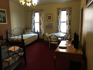 Ein Zimmer mit einem Bett, einem Schreibtisch und einem Stuhl. Es gibt ein Fenster mit Vorhängen, ein Bild an der Wand und eine Deckenleuchte.