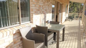 A covered outdoor seating area with wicker furniture and a glass table, adjacent to a brick wall and large windows. - Pacific Breeze - ocean views from BBQ deck (DUNBOGAN)