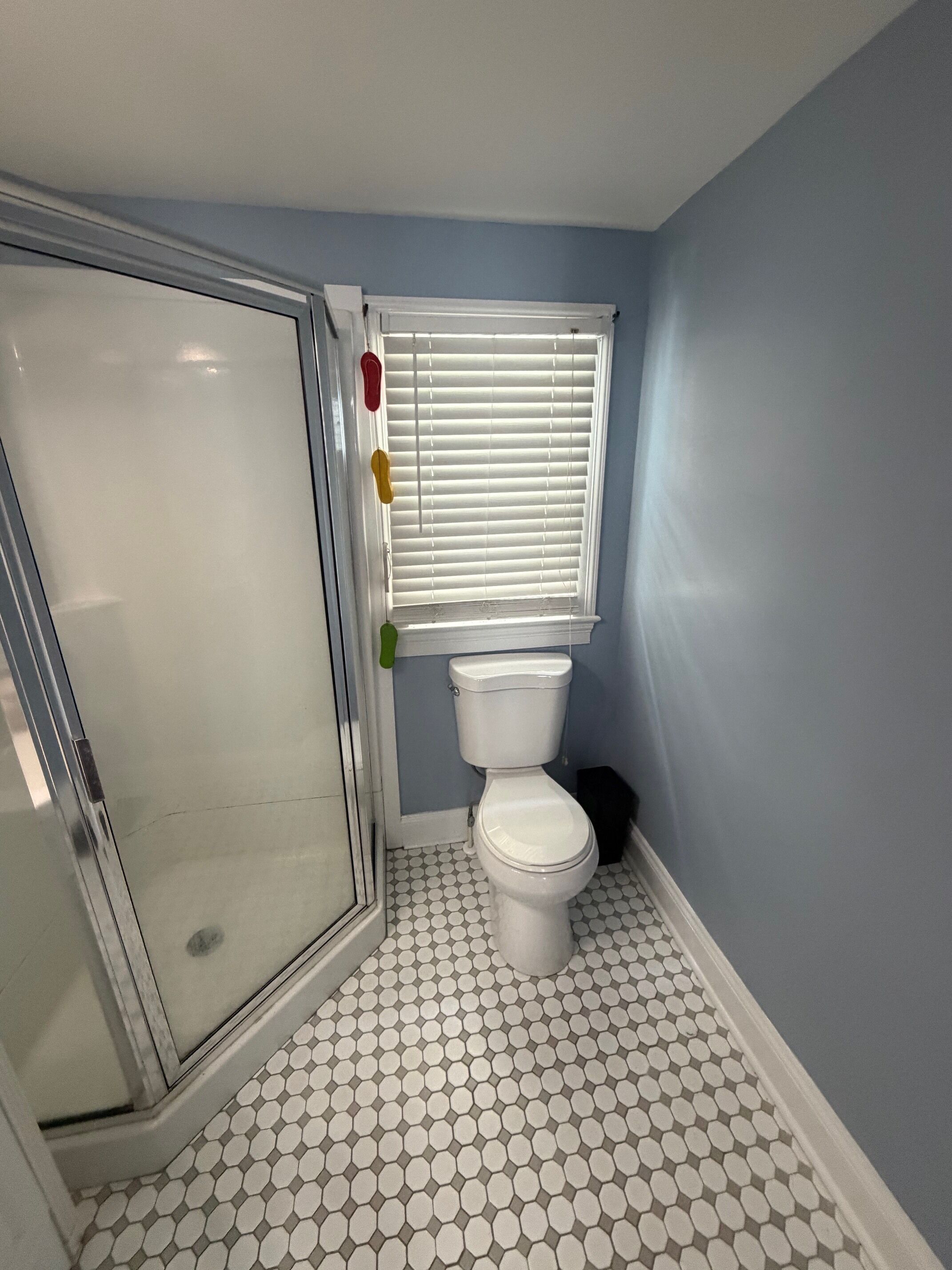 Une salle de bain avec des toilettes blanches, une douche dotée d’une porte en verre et une fenêtre avec des stores.
