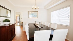 Een eetkamer met een donker houten tafel, witte stoelen, een kroonluchter, een lijstwerk schilderij en een raam met jaloezieën.
