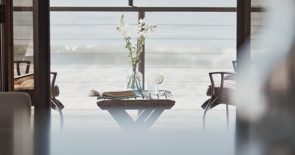 A modern room with a large window, a small table with a book and a vase of flowers, and two chairs. - Beachfront Retreat On Sand with Charming Nautical Features | Lower Unit (Santa Barbara)