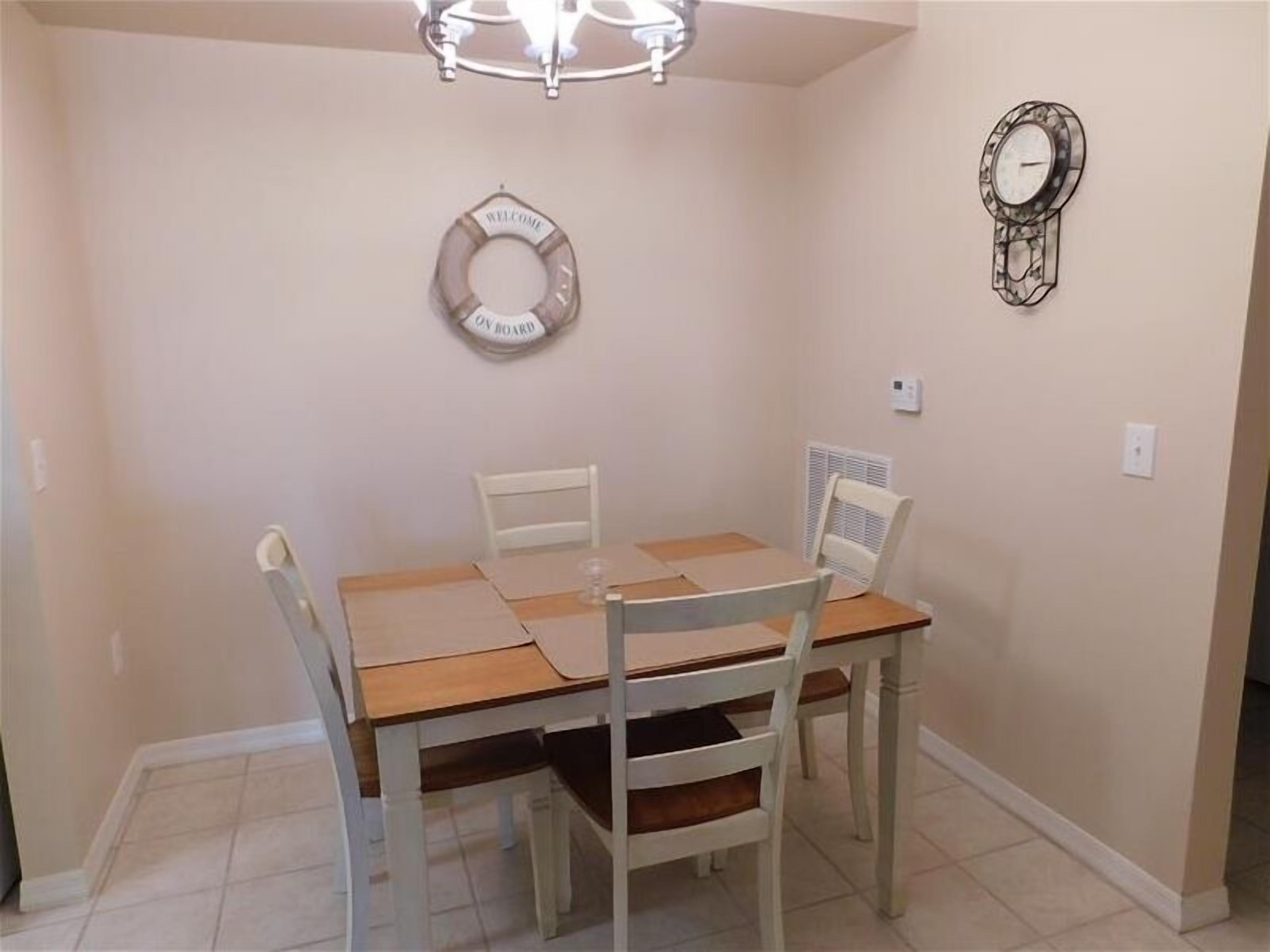 Uma sala de jantar com mesa de madeira e quatro cadeiras, um candelabro, um letreiro de boas-vindas e um relógio na parede.