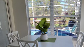 Ein Essbereich mit weißem Tisch und Stühlen, einer Topfpflanze und Blick auf einen Außenpool durch große Fenster.