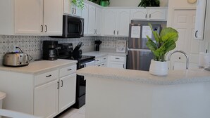 Eine moderne Küche mit weißen Schränken, einem Edelstahlkühlschrank, einer Mikrowelle und einem Toaster. Auf den Arbeitsflächen stehen Pflanzen und an der Wand hängt eine Uhr.