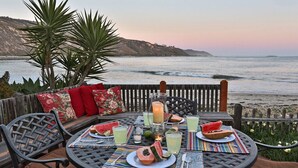 A dining table set with plates, cutlery, and drinks, overlooking a beach with waves and hills in the background. - Rincon Retreat - Casual Beachfront Comfort (Carpinteria)