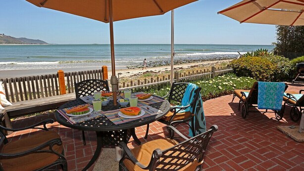 An outdoor patio with a table set for dining, overlooking a beach. - Rincon Retreat - Casual Beachfront Comfort (Carpinteria)