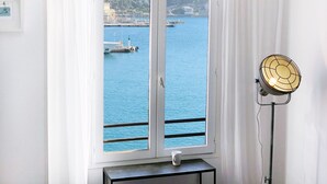 A room with a window overlooking a harbor, a white curtain, a table with a cup, and a standing fan. - Waterfront panoramic sea view with balcony, AC (Nice)