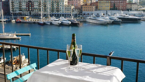 A table with a bottle of champagne and two glasses, overlooking a marina with yachts and colorful buildings in the background. - Waterfront panoramic sea view with balcony, AC (Nice)