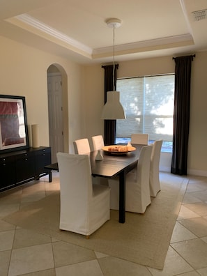 Un comedor con una mesa, sillas, una lámpara colgante y vistas al exterior a través de una ventana con cortinas.
