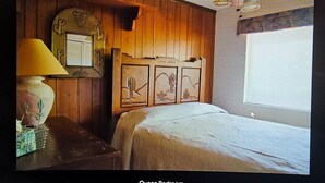Un dormitorio con paneles de madera, una cama con cubrecama blanco, una mesita de noche con una lámpara y un reloj fijado en la pared.