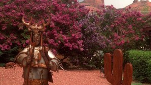 Una escultura de una figura con un báculo y un cactus en un jardín con grava roja y flores púrpuras.