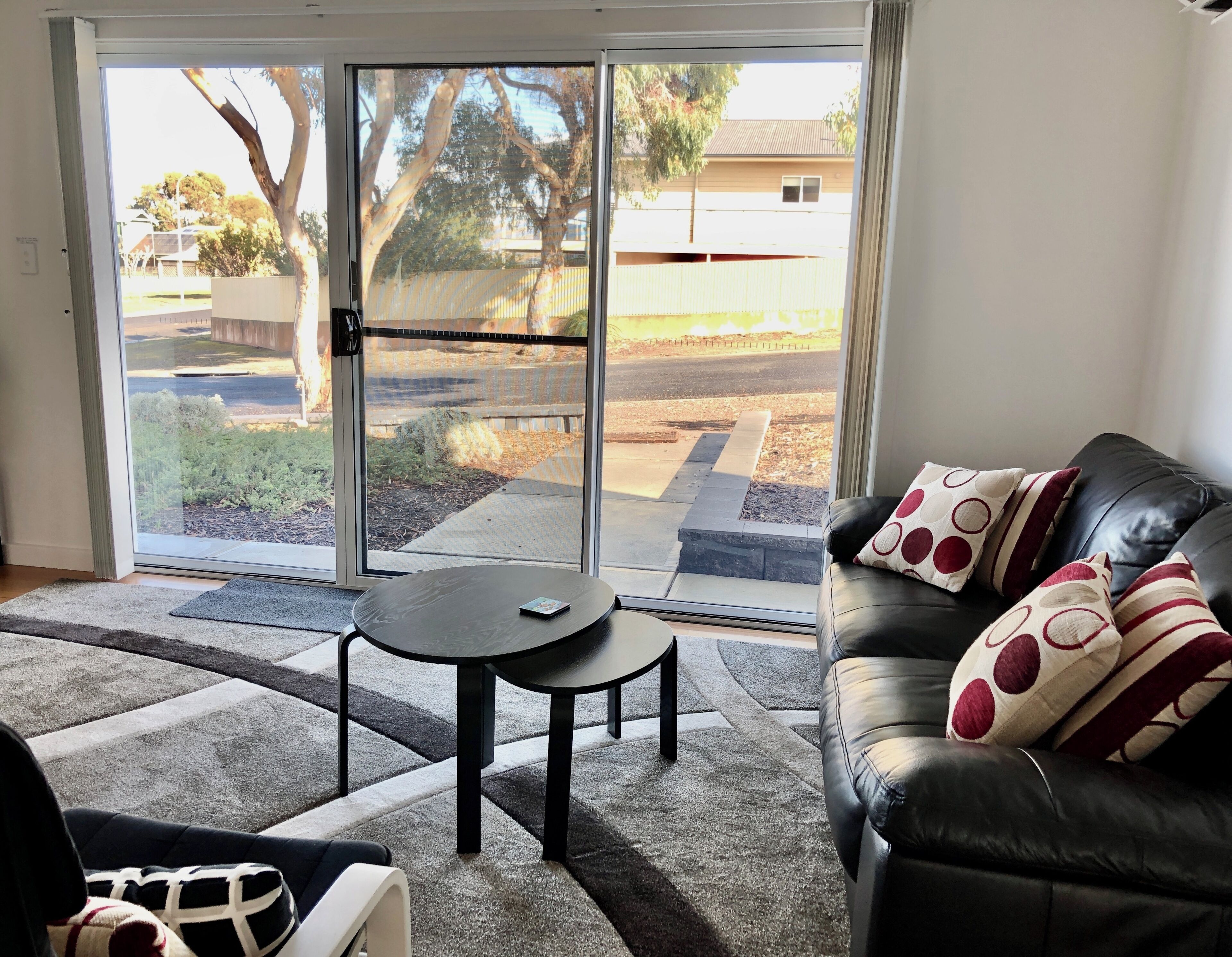 A living room with a black leather sofa, a round table, and a sliding glass door leading to a backyard.