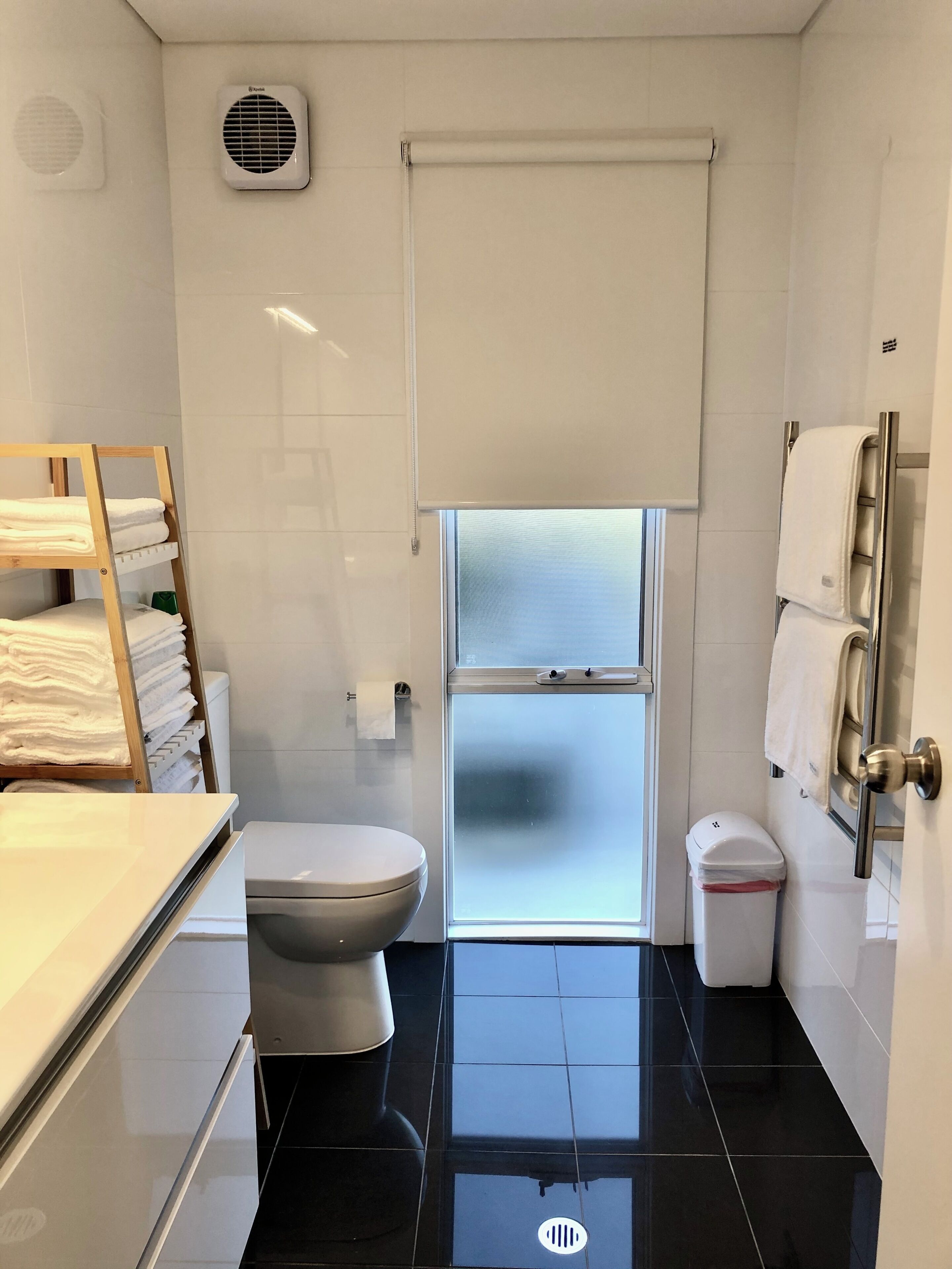 A modern bathroom with a toilet, a towel rack, a small trash bin, and a window with frosted glass.