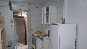 Een compacte keuken met witte kasten, een koelkast, een fornuis en een spoelbak. Er staat een stoel en een klein tafeltje met een mandje erop.