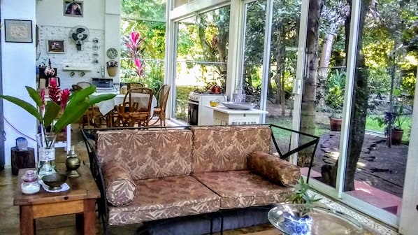 Una habitación acogedora con un sofá estampado, una mesa de comedor con sillas y una cocina visible a través de amplios ventanales.