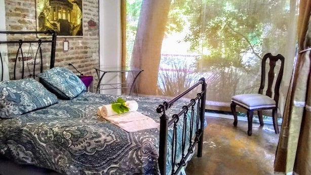 Un dormitorio con una colcha estampada, una mesita con una silla y vistas a árboles por la ventana.