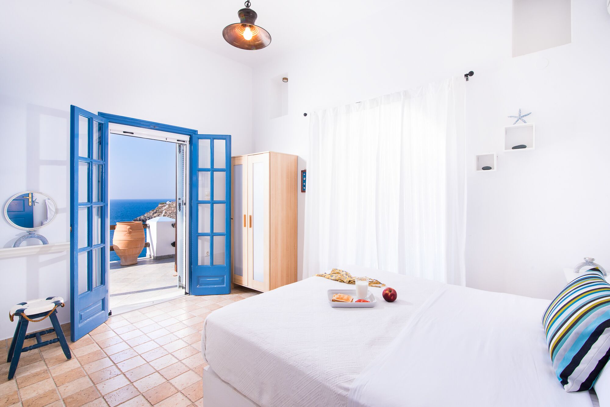 A bedroom with a bed, a bedside table, a wardrobe, a window with curtains, and a view of the sea.