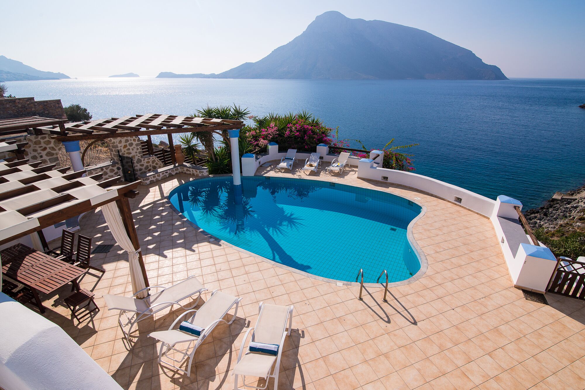 A pool area with a view of the sea and a mountain, surrounded by lounge chairs and a pergola.